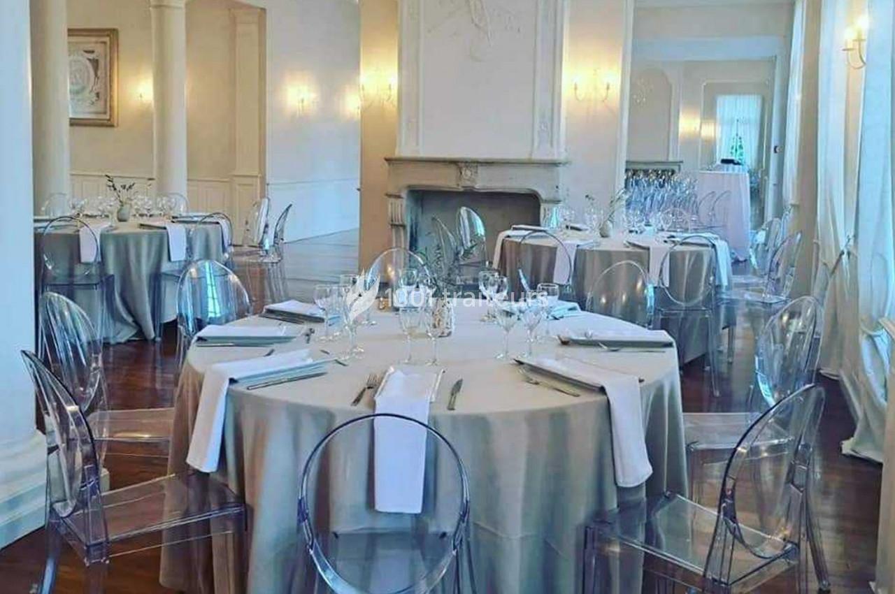 Salle élégante avec tables rondes dressées, nappes beiges, chaises transparentes et lumière naturelle.
