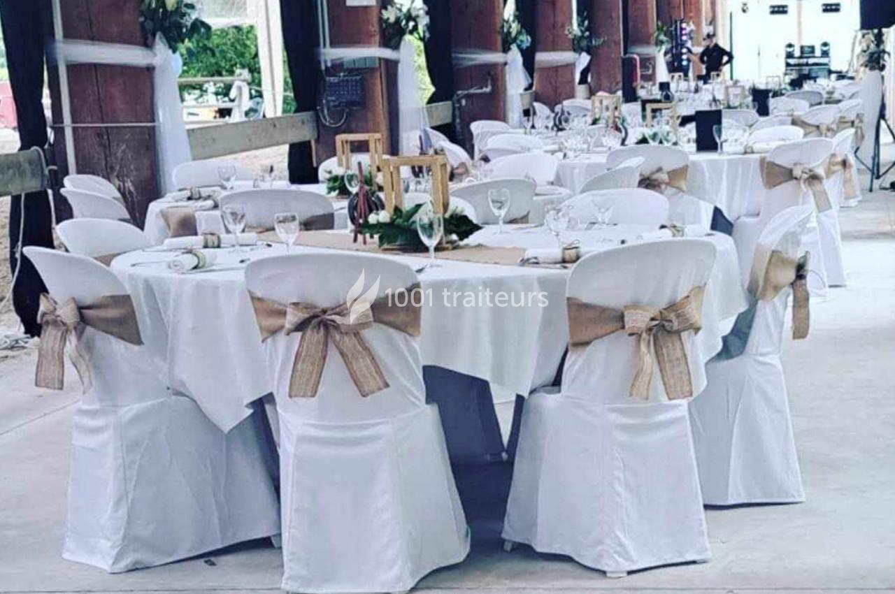Salle décorée pour un événement avec des tables rondes dressées, nappes blanches et chaises ornées de nœuds en toile de jute.