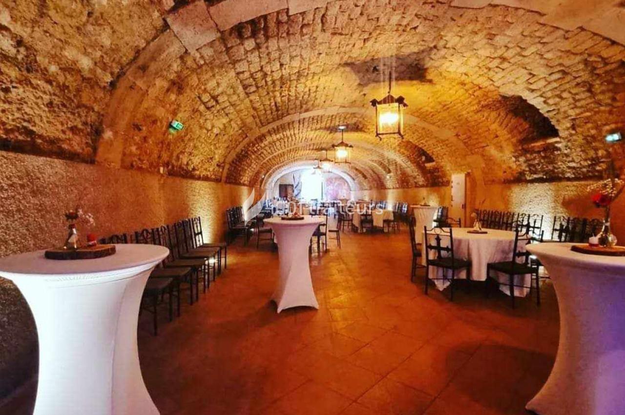 Salle voûtée en pierre avec tables rondes dressées et mange-debout, éclairée par des lanternes suspendues.