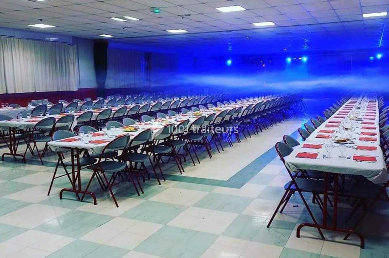 Salle de réception avec de longues tables dressées, nappes blanches, serviettes rouges et éclairage bleu tamisé.