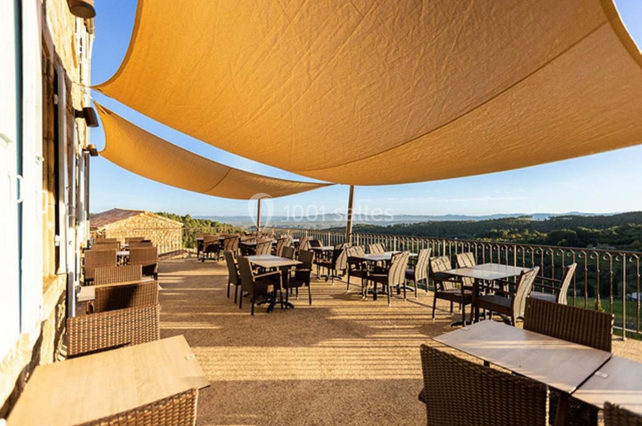 Terrasse ensoleillée avec tables et chaises en rotin, ombragée par de grandes toiles tendues, vue sur la campagne.