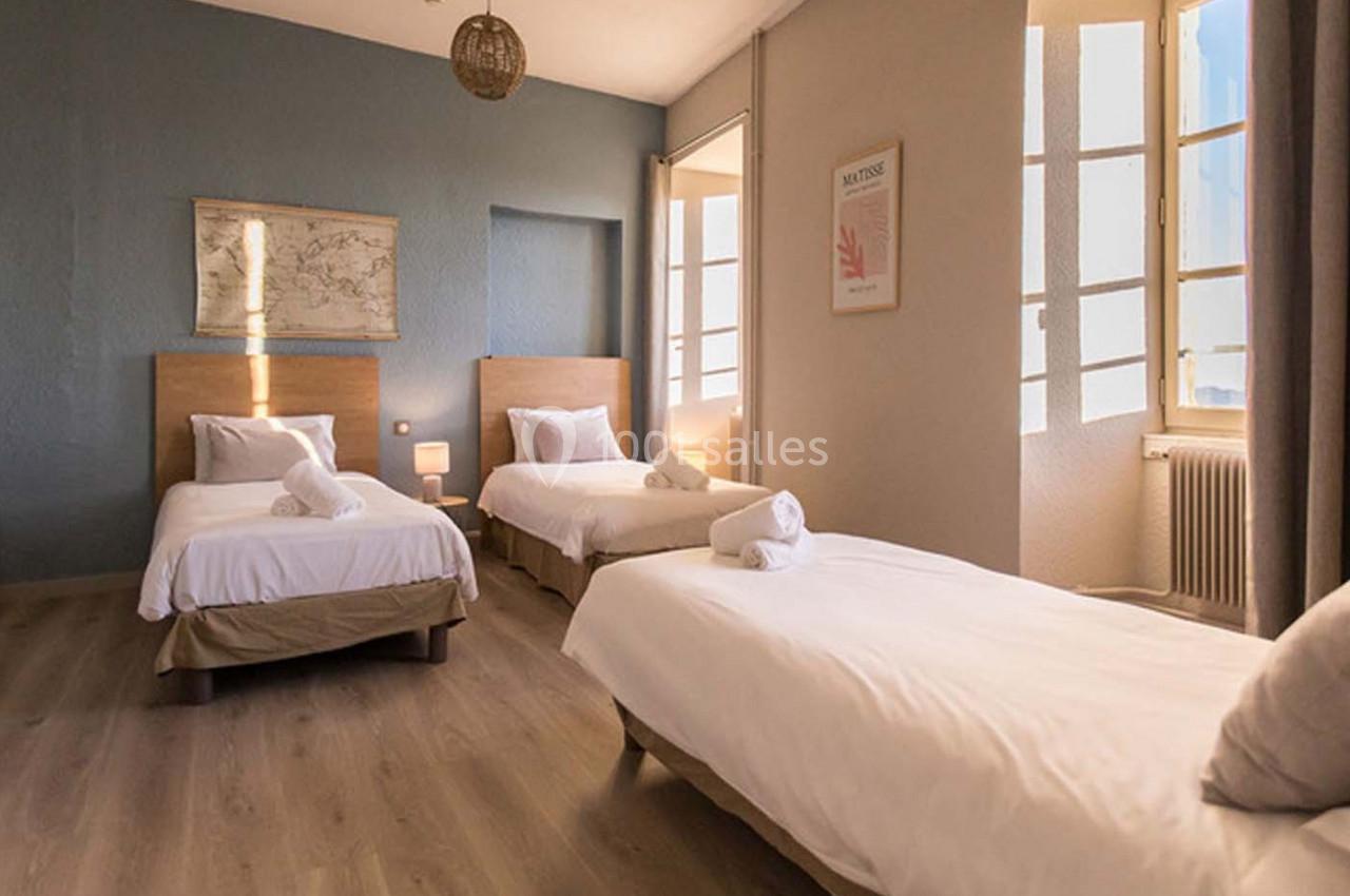 Chambre lumineuse avec trois lits simples, parquet au sol, murs clairs et grandes fenêtres laissant entrer la lumière…