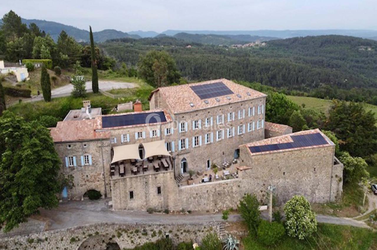 Vue aérienne d'un grand bâtiment en pierre avec des panneaux solaires sur le toit, entouré de collines verdoyantes.