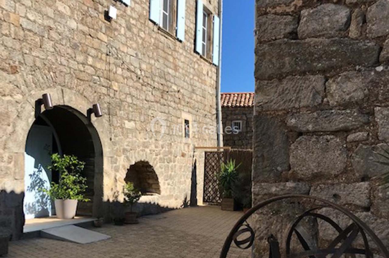 Cour pavée entourée de murs en pierre, avec des plantes en pot et une porte voûtée blanche.