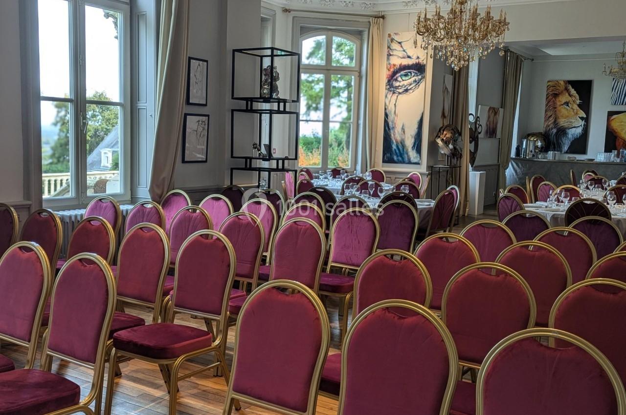 Salle de réception avec des chaises rouges alignées, grandes fenêtres, lustre et tableaux décoratifs au mur.