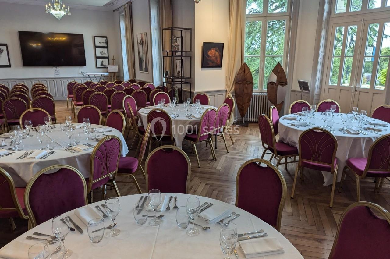 Salle élégante avec tables rondes dressées, chaises rouges, grandes fenêtres et décorations murales.