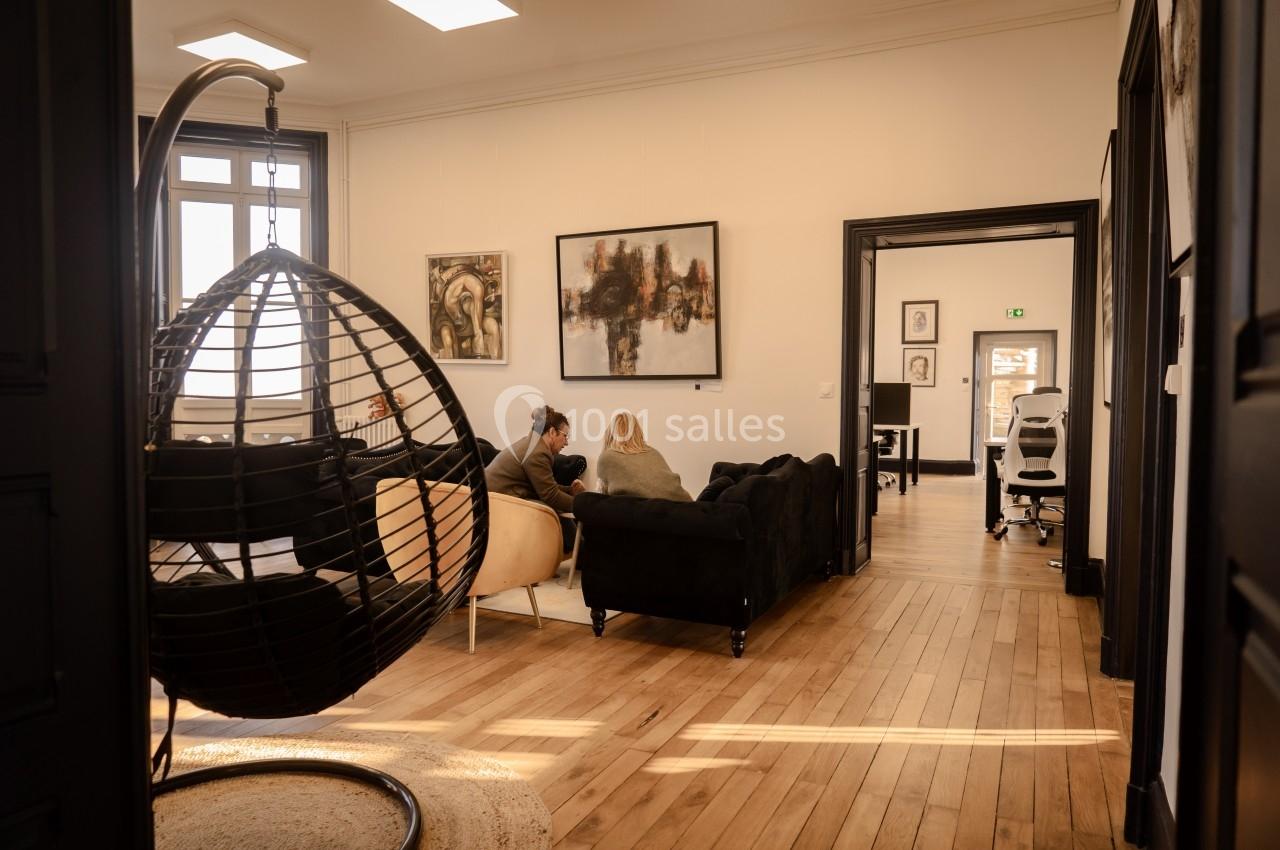 Deux personnes discutent sur des canapés dans un salon lumineux avec parquet, fauteuil suspendu et tableaux au mur.