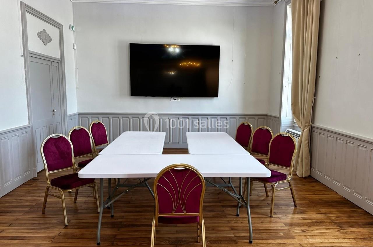 Salle lumineuse avec parquet, table blanche rectangulaire et chaises rouges disposées autour, écran mural en arrière-plan.