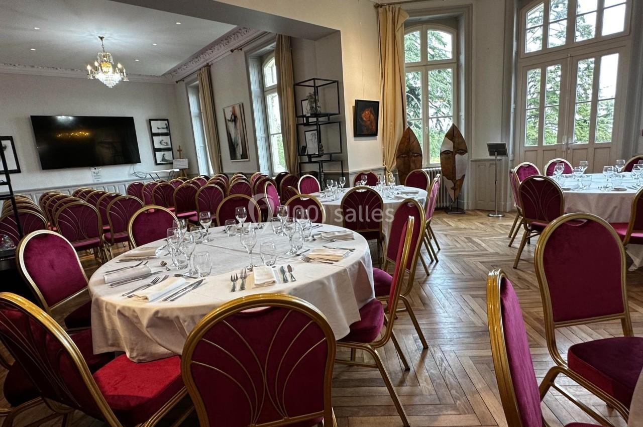 Salle élégante avec tables rondes dressées, chaises rouges, parquet en bois et grandes fenêtres lumineuses.