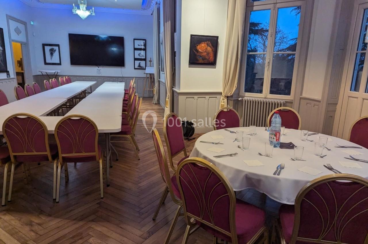 Salle de réunion avec tables rectangulaires et rondes, chaises rouges, parquet en bois et fenêtres donnant sur l'extérieur.