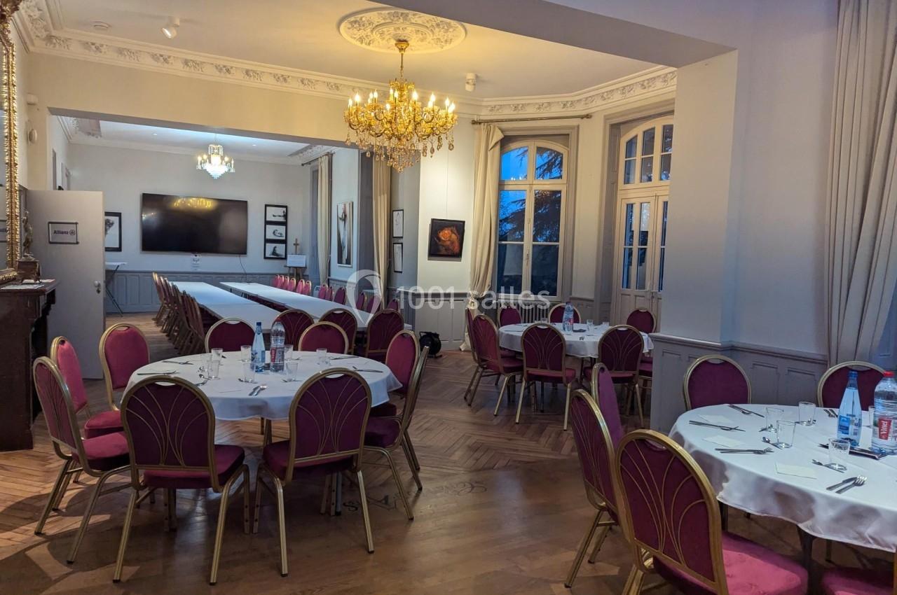 Salle élégante avec tables rondes dressées, chaises rouges, parquet en bois, lustre doré et grandes fenêtres.