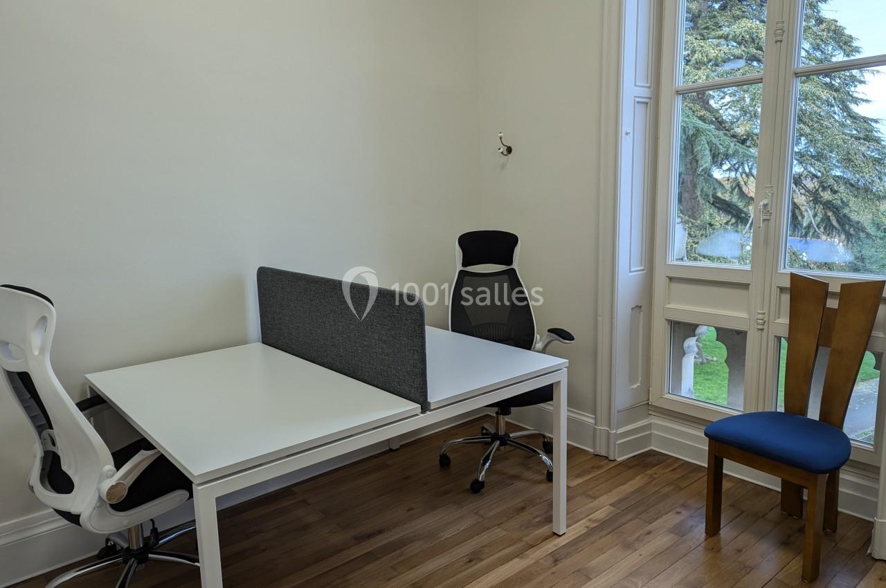 Deux bureaux blancs avec chaises, séparés par une cloison grise, près d'une fenêtre donnant sur un jardin.