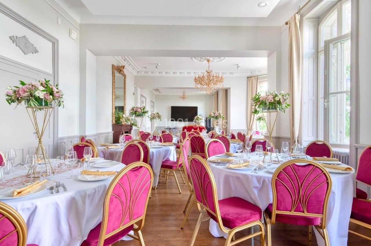 Salle de réception élégante avec tables dressées, chaises roses, nappes blanches et décorations florales.
