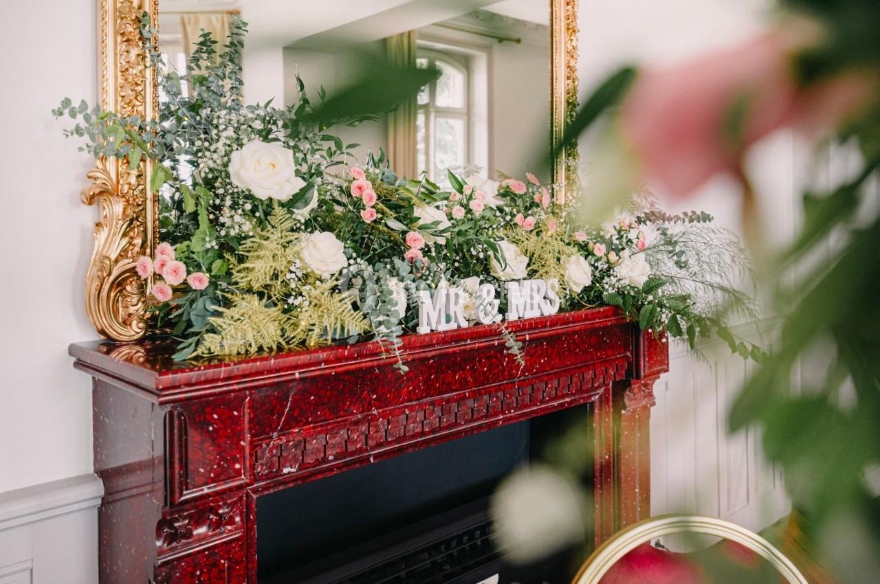 Cheminée rouge ornée de fleurs blanches et roses, avec des lettres ’MR & MRS’ posées devant un miroir doré.