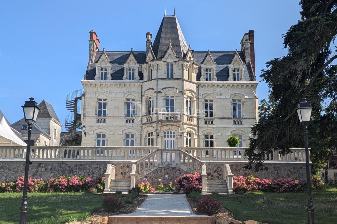 Façade d'un château en pierre claire avec escaliers, jardin fleuri et lampadaires, sous un ciel bleu.