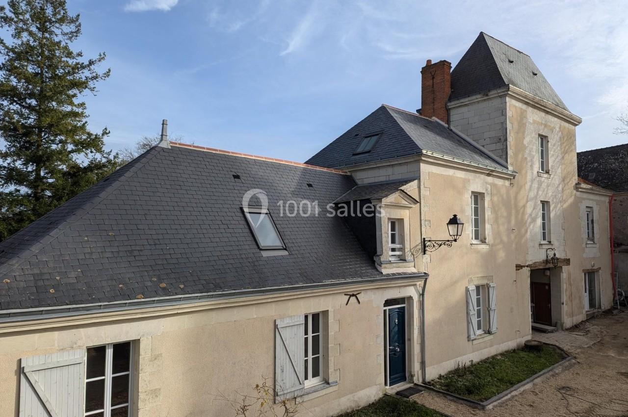 Façade d'une maison en pierre avec toiture en ardoise, fenêtres à volets et une tour attenante sous un ciel dégagé.