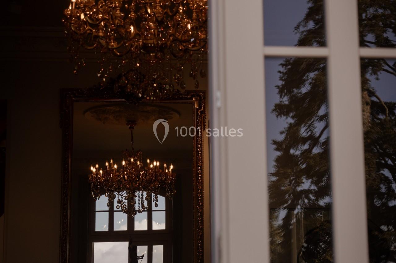 Reflet d'un lustre doré et d'une fenêtre dans un miroir, avec un arbre visible à travers une porte entrouverte.