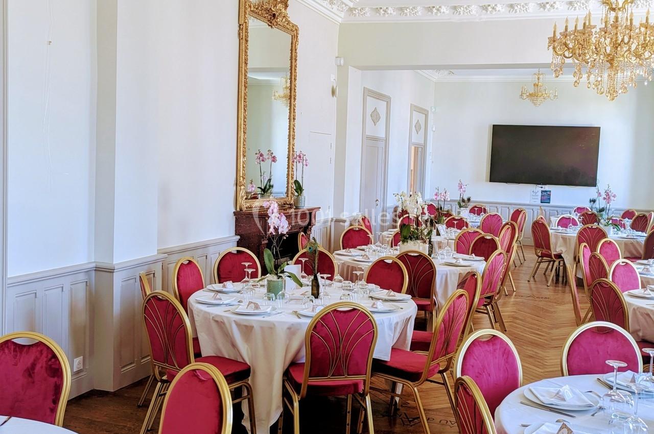 Salle de réception élégante avec tables dressées, chaises rouges, lustres dorés et grand miroir mural.