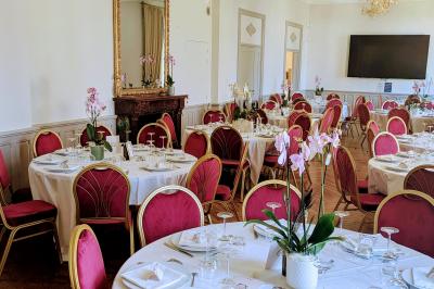 Salle élégante avec tables rondes dressées, chaises rouges et dorées, grand miroir doré et orchidées en décoration.