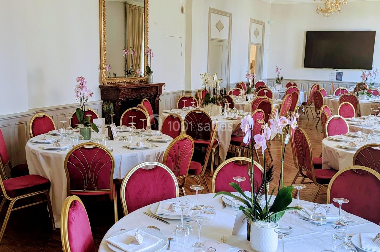 Salle de réception élégante avec tables rondes dressées, chaises rouges, miroir doré et lustre en cristal.