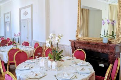 Salle élégante avec tables rondes dressées, chaises rouges et dorées, grand miroir doré et orchidées en décoration.