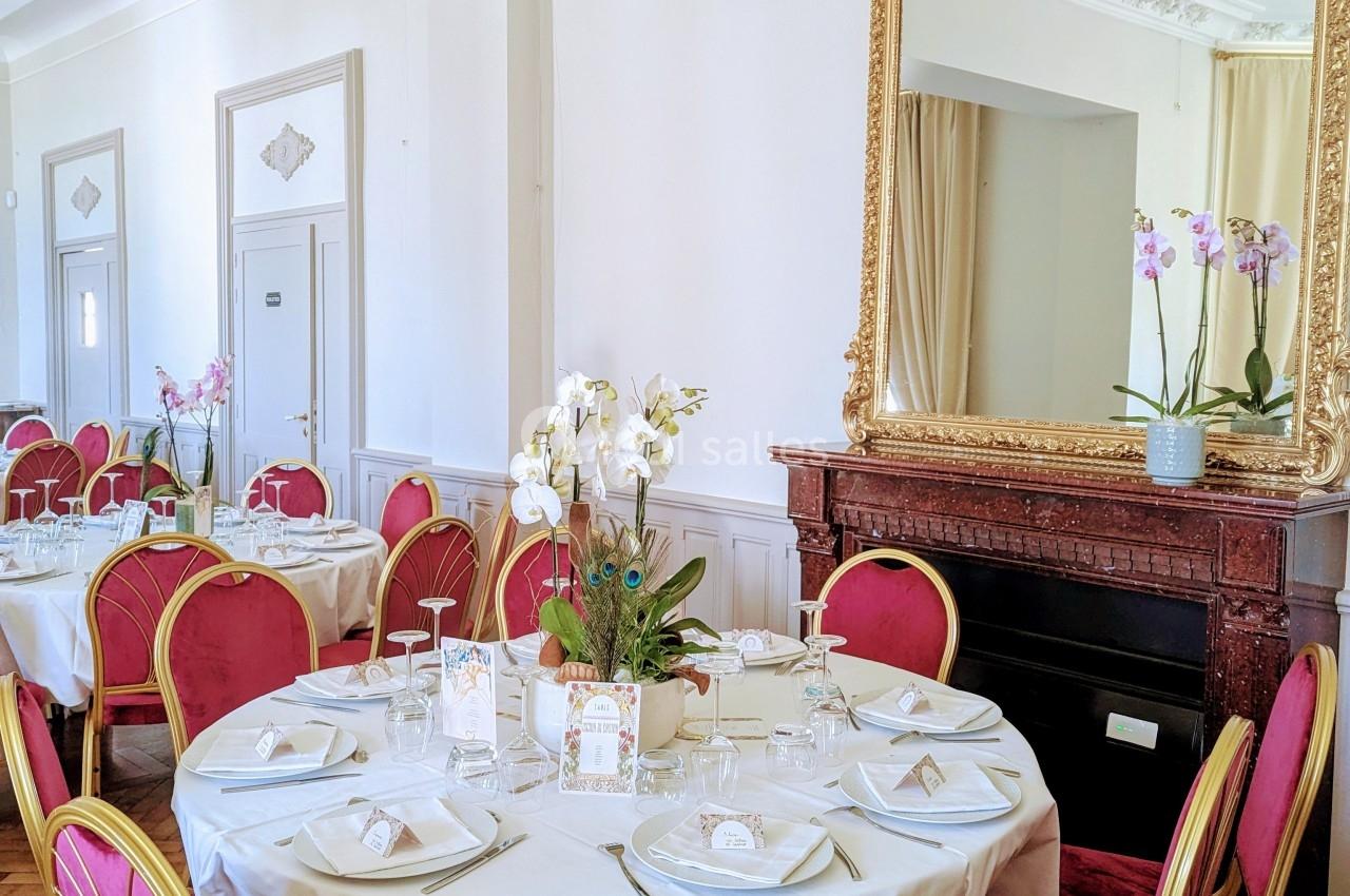 Salle élégante avec tables rondes dressées, chaises rouges et dorées, grand miroir doré et orchidées en décoration.