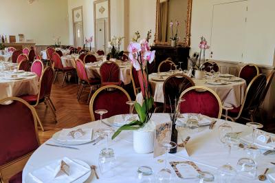 Salle élégante avec tables rondes dressées, chaises rouges et dorées, grand miroir doré et orchidées en décoration.