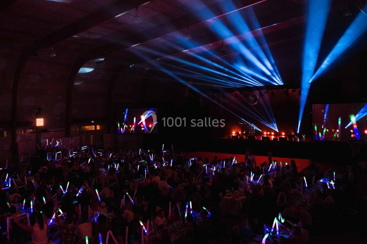 Salle sombre remplie de spectateurs tenant des bâtons lumineux, éclairée par des projecteurs et des faisceaux lumineux…