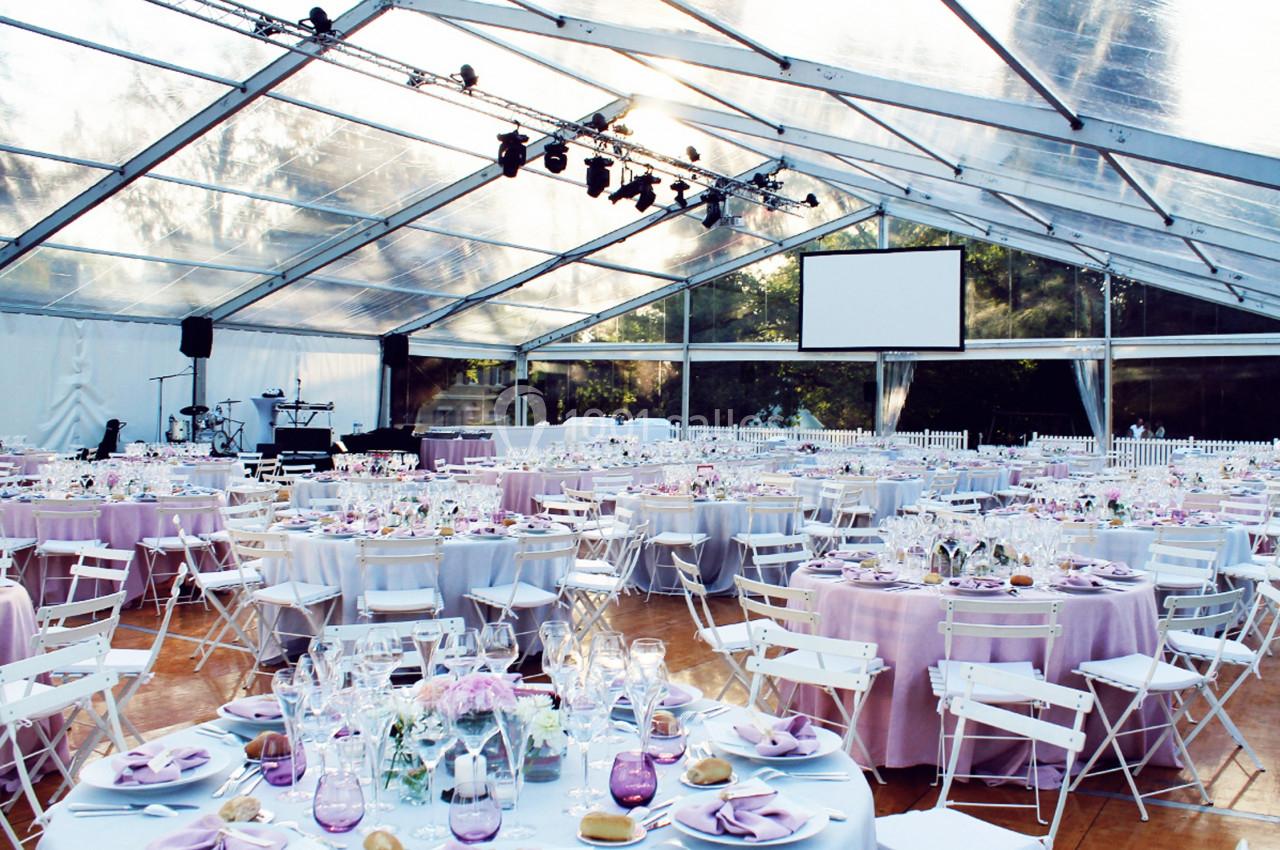 Salle de réception sous une tente transparente, avec tables rondes dressées et décorations dans des tons pastel.