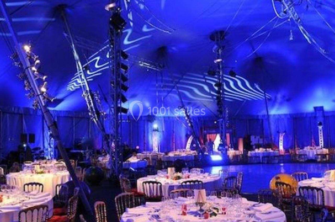 Salle de réception sous un chapiteau éclairé en bleu, avec des tables rondes dressées pour un événement.