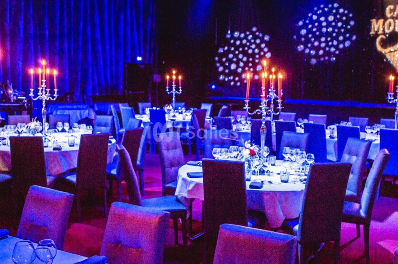 Salle de cabaret avec tables dressées, chandeliers allumés et éclairage tamisé, décorée pour un spectacle.