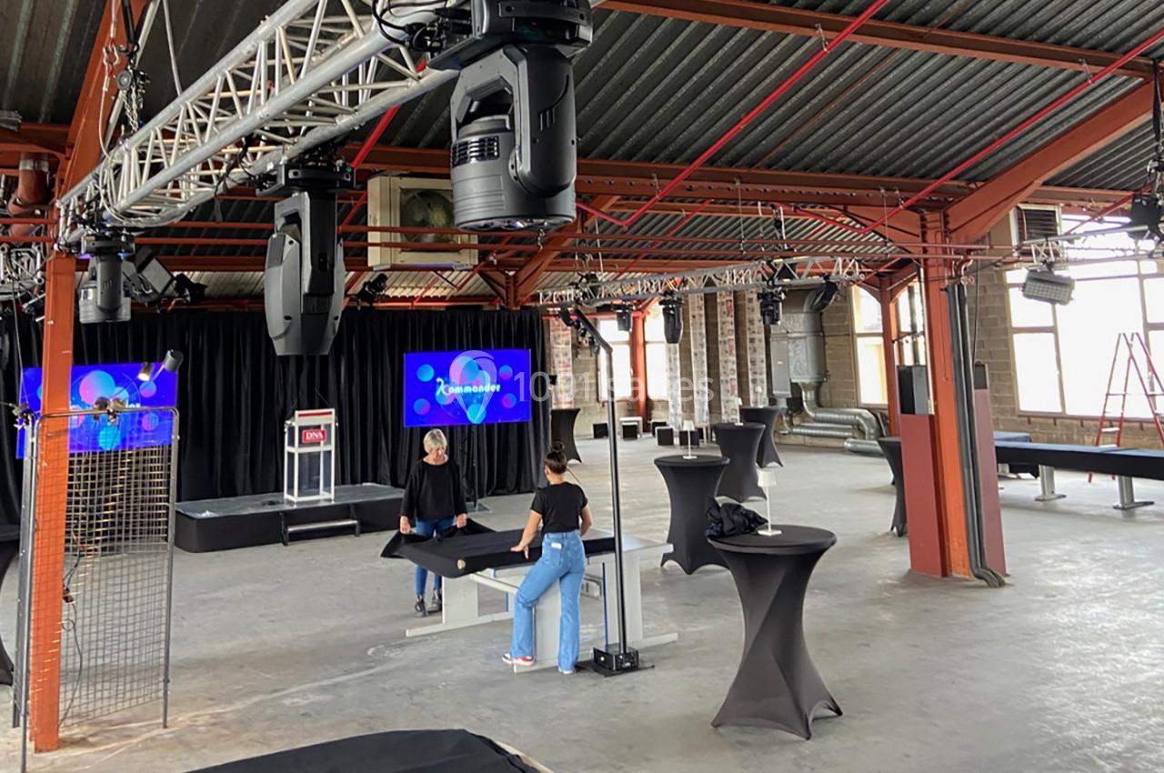 Deux personnes installent des tables dans une grande salle industrielle équipée de projecteurs et d'écrans.