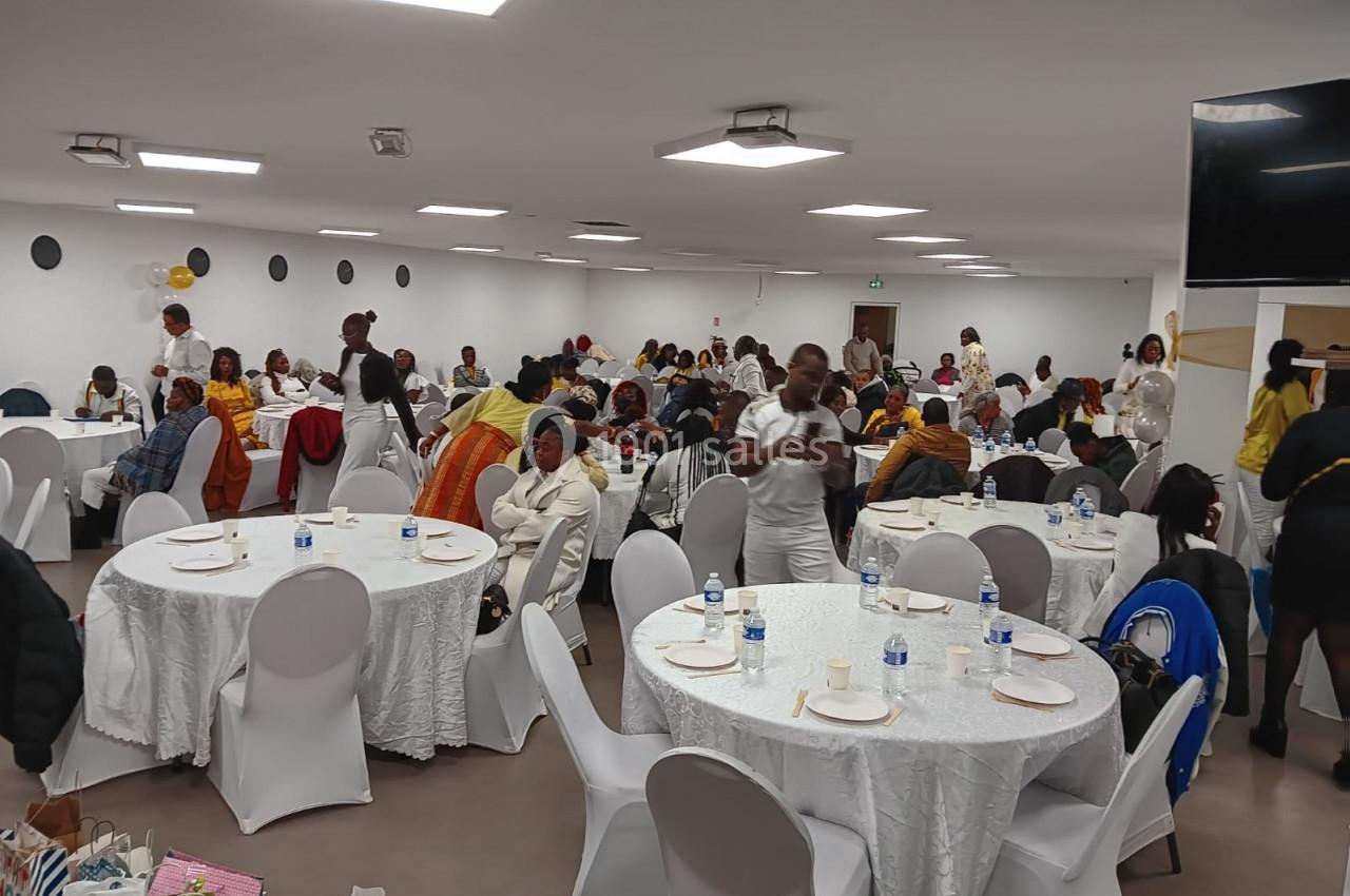 Des personnes assises et debout dans une grande salle lumineuse, aménagée avec des tables rondes et des chaises blanches.