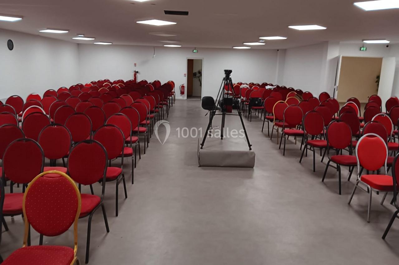 Salle de conférence vide avec des rangées de chaises rouges et un trépied de caméra au centre.