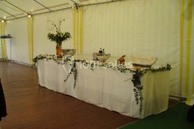 Tente avec table dressée pour un buffet, entourée de chaises blanches, installée sur une pelouse avec haies en arrière-plan.