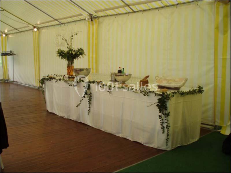 Table décorée avec nappes blanches, feuillage et vaisselle, installée sous une tente à rayures jaunes et blanches.
