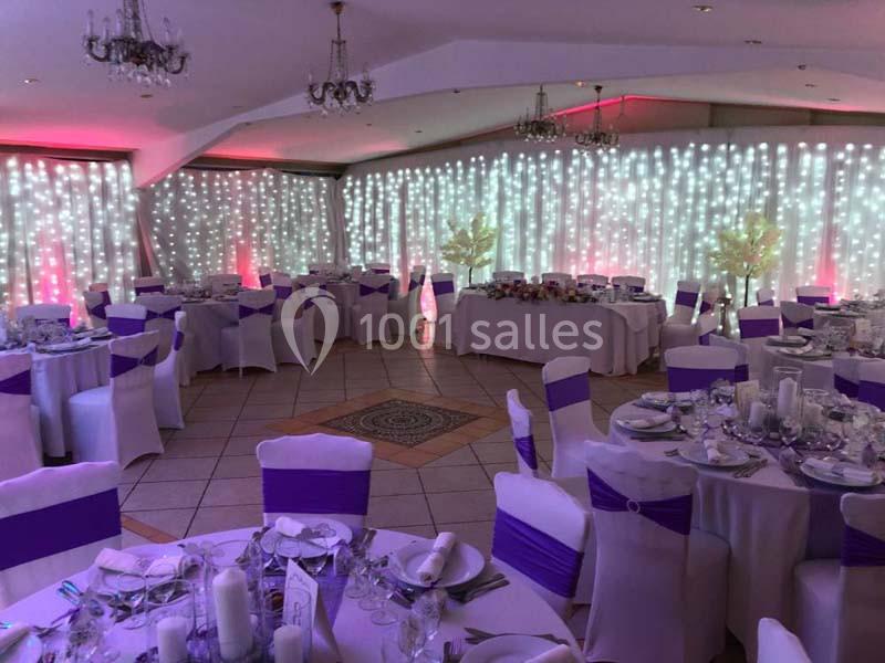 Salle de réception décorée avec des nappes blanches, des chaises ornées de rubans violets et un fond lumineux.