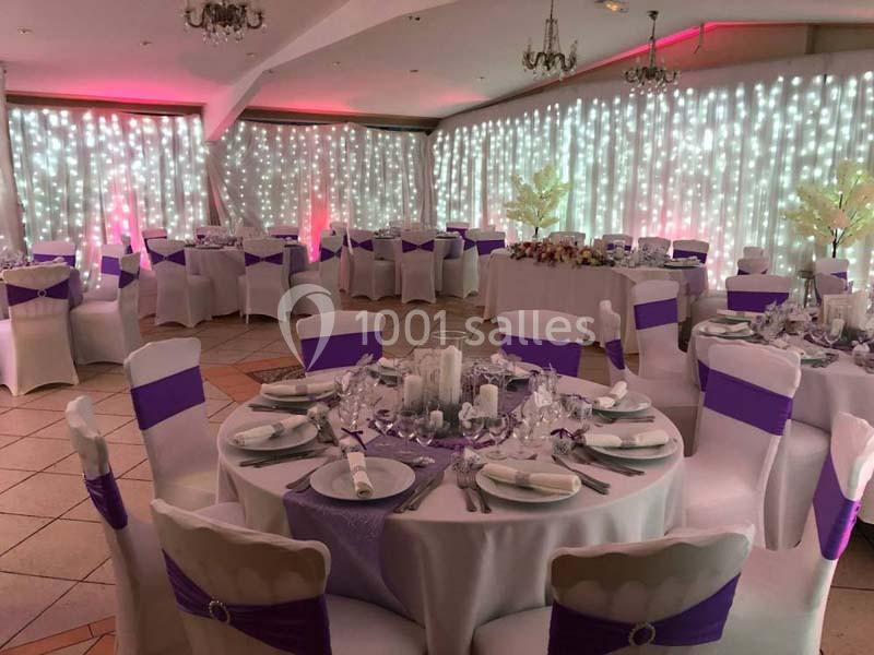 Salle de réception décorée avec des tables rondes, nappes blanches, chemins de table violets et guirlandes lumineuses.