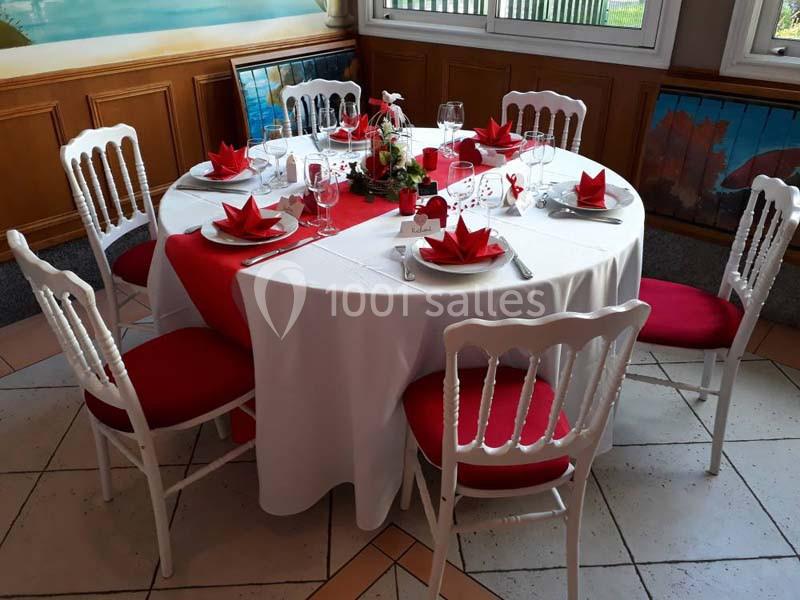 Table ronde dressée avec une nappe blanche, serviettes pliées en rouge, chaises assorties et décoration centrale florale.