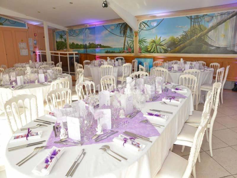 Salle de réception décorée avec des tables rondes dressées, nappes blanches et accents violets, fresque murale tropicale.
