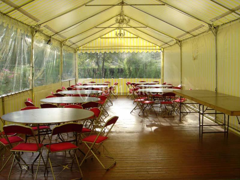 Intérieur d'une tente avec des tables rondes et des chaises rouges disposées sur un sol en bois, éclairée par la lumière…