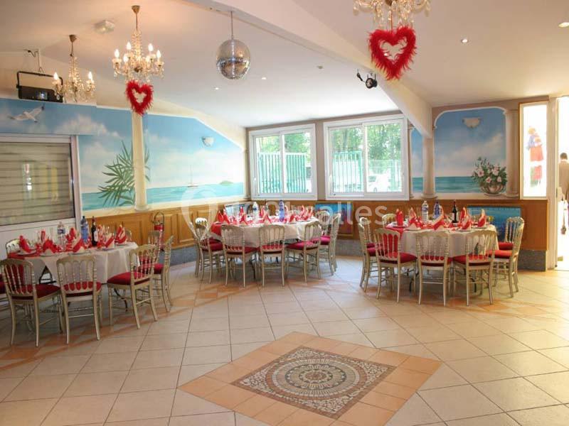 Salle décorée avec des tables dressées, nappes blanches, chaises rouges et cœurs suspendus, ambiance festive.