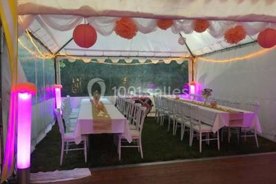 Tente avec table dressée pour un buffet, entourée de chaises blanches, installée sur une pelouse avec haies en arrière-plan.