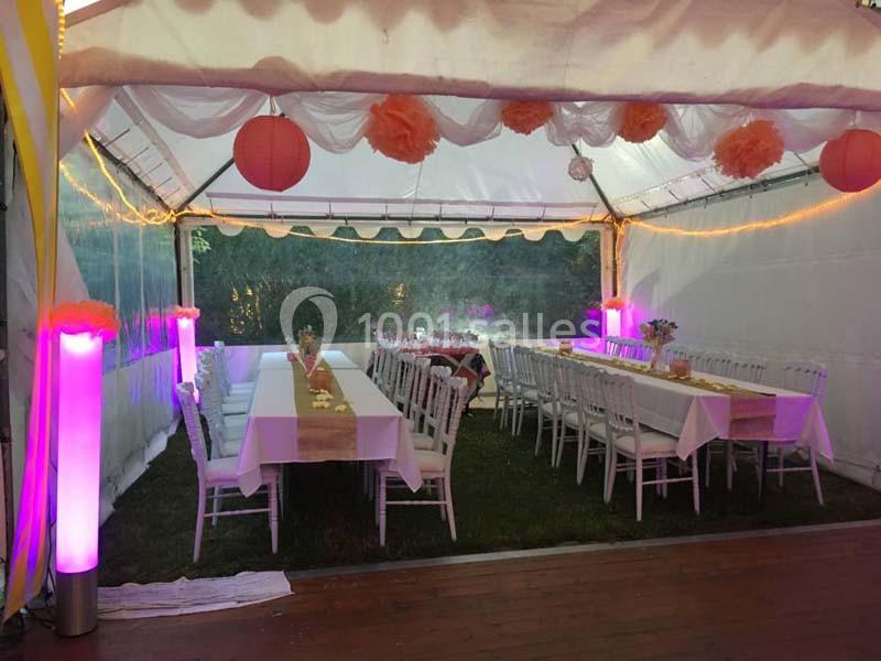 Intérieur d'une tente décorée pour un événement, avec tables dressées, chaises blanches et éclairage coloré.