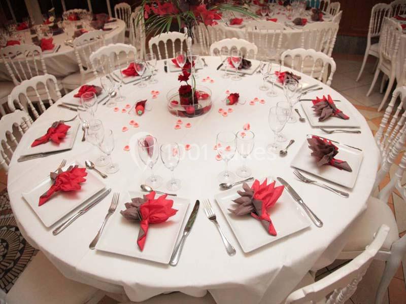 Table ronde dressée pour un événement, avec nappes blanches, serviettes pliées en fleurs rouges et violettes, et vaisselle…