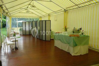 Tente avec table dressée pour un buffet, entourée de chaises blanches, installée sur une pelouse avec haies en arrière-plan.