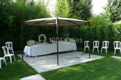 Tente avec table dressée pour un buffet, entourée de chaises blanches, installée sur une pelouse avec haies en arrière-plan.