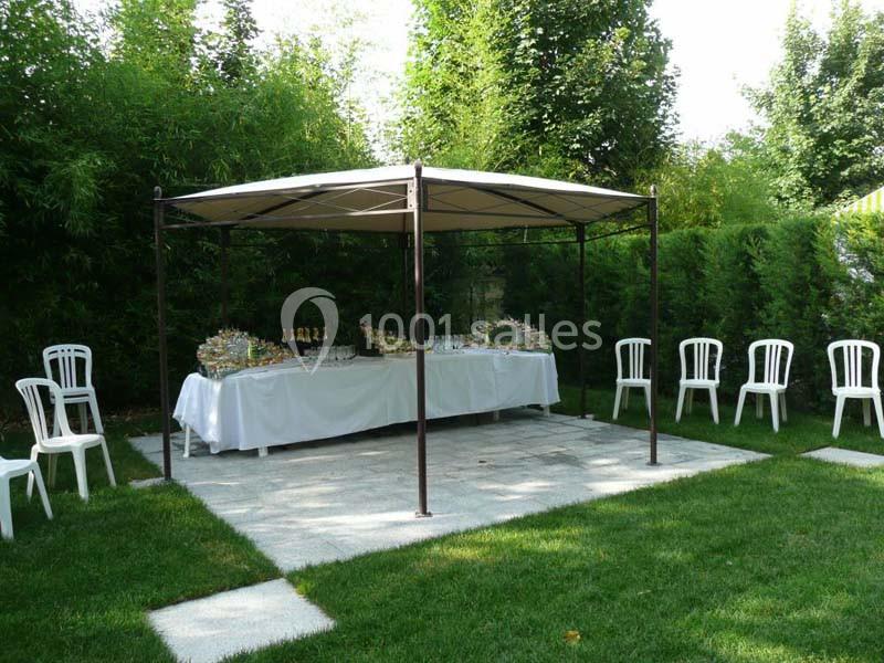 Tente avec table dressée pour un buffet, entourée de chaises blanches, installée sur une pelouse avec haies en arrière-plan.