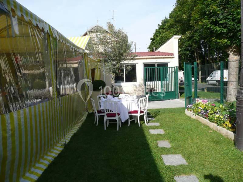Table dressée dans un jardin, entourée de chaises blanches, près d'une tente rayée jaune et blanche et d'un bâtiment.