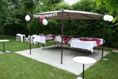 Tente avec table dressée pour un buffet, entourée de chaises blanches, installée sur une pelouse avec haies en arrière-plan.